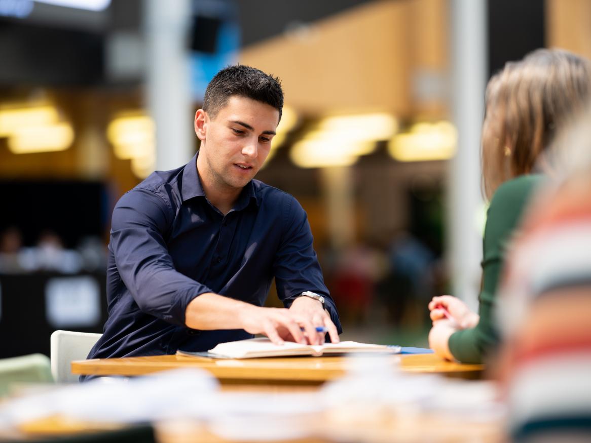 Male postgraduate student sitting at a desk with a colleague referring to an open book