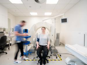 Female research study participant seated in front of gamma camera, researcher out of focus