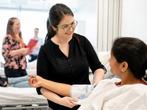 Female research study participant having blood pressure checked by researcher, in three-bay bed room. Other participants and researchers out of focus in the background