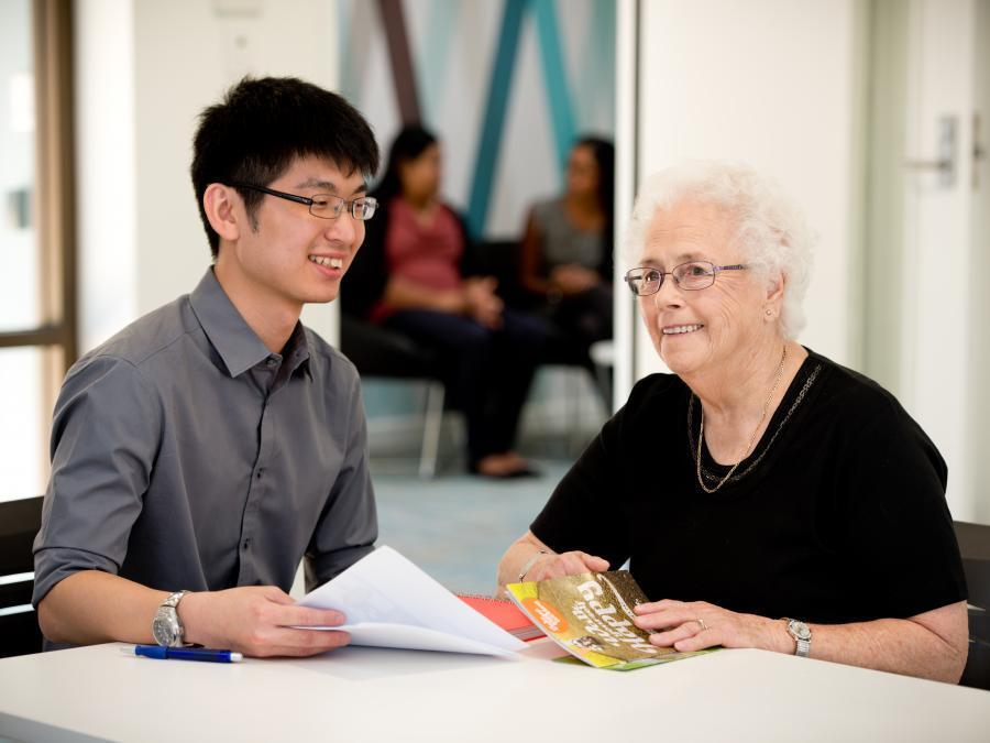 Young man sitting at a desk with elderly woman, discussing paperwork