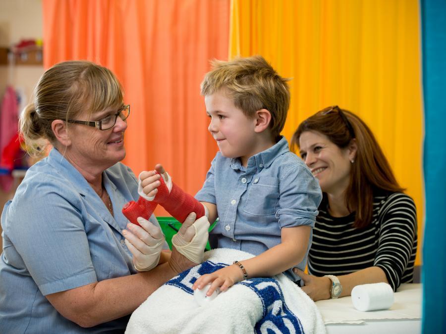 Child sitting up in hospital bed with red plaster cast on arm, smiling at mum and nurse