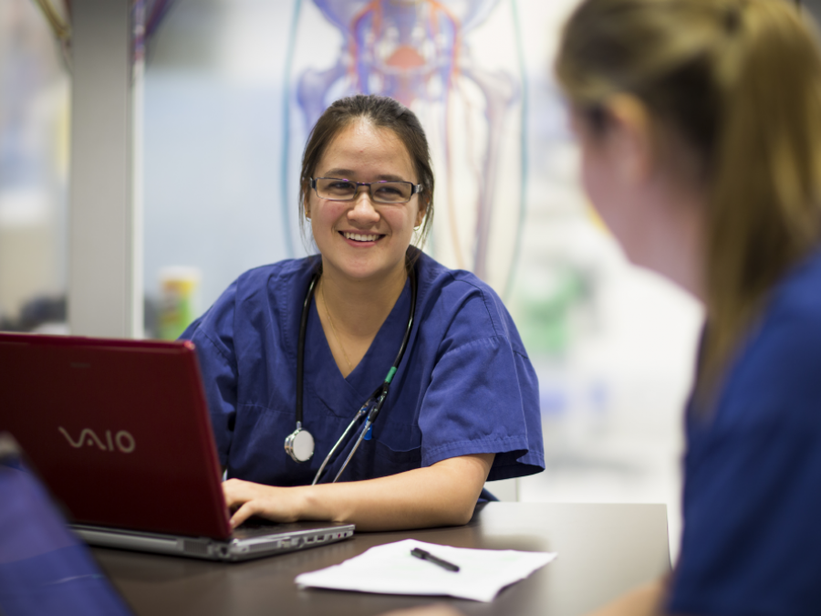Female doctor in scrubs sitting in front of a laptop at a desk, smiling to colleague
