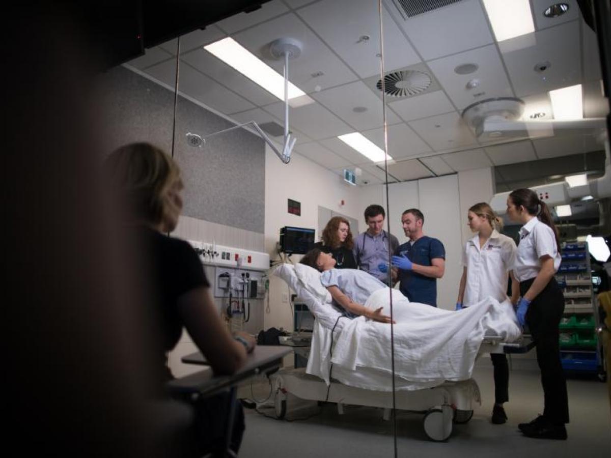 Adam Montagu talking to group of students in simulated hospital setting while assessor watches from darkened room