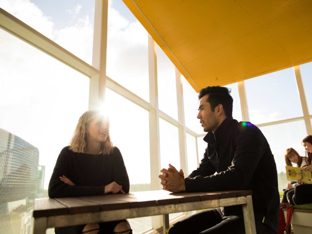 Two students sitting at a table inside the AHMS building talking, with sun flare through windows behind them