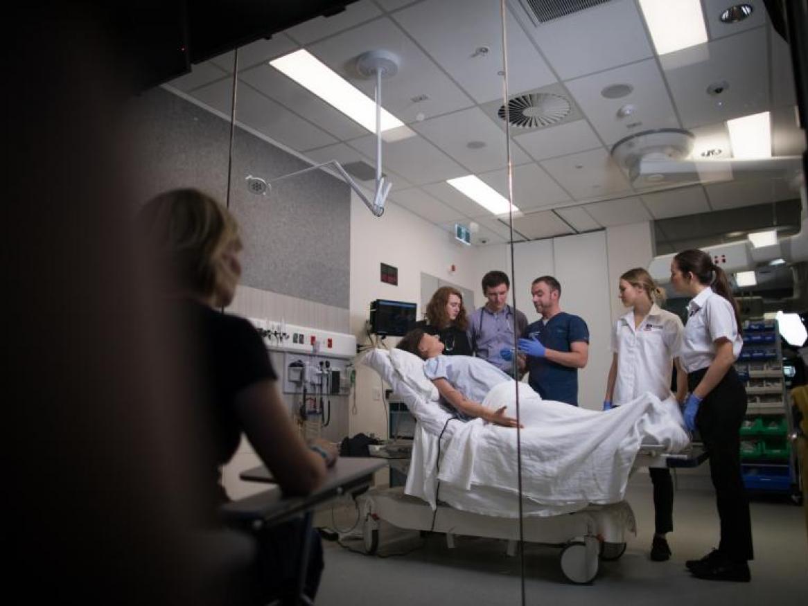 Adam Montagu talking to group of students in simulated hospital setting while assessor watches from darkened room