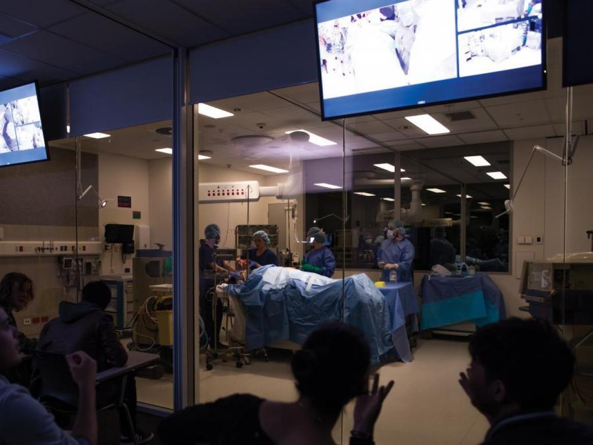 students in simulated operating room, watched by onlookers through glass observation windows