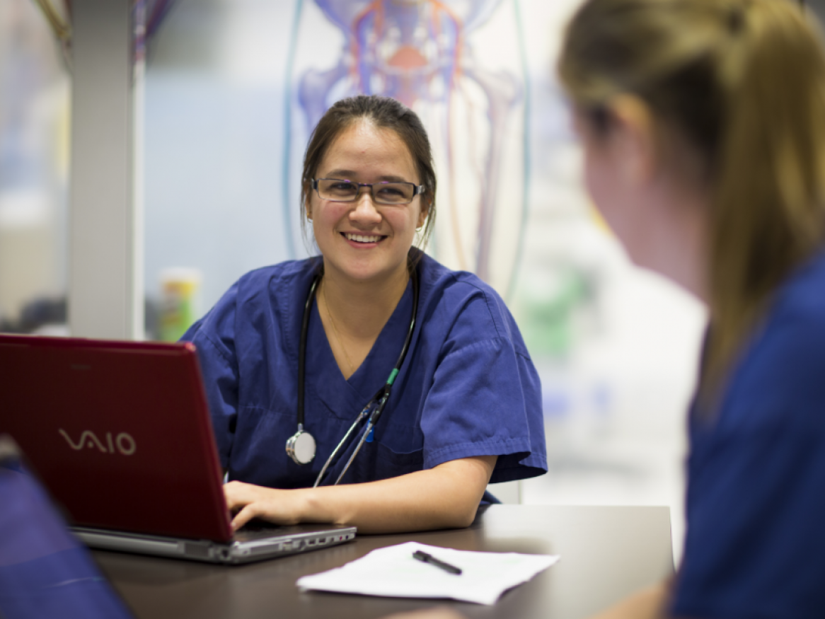 Female doctor in scrubs sitting in front of a laptop at a desk, smiling to colleague