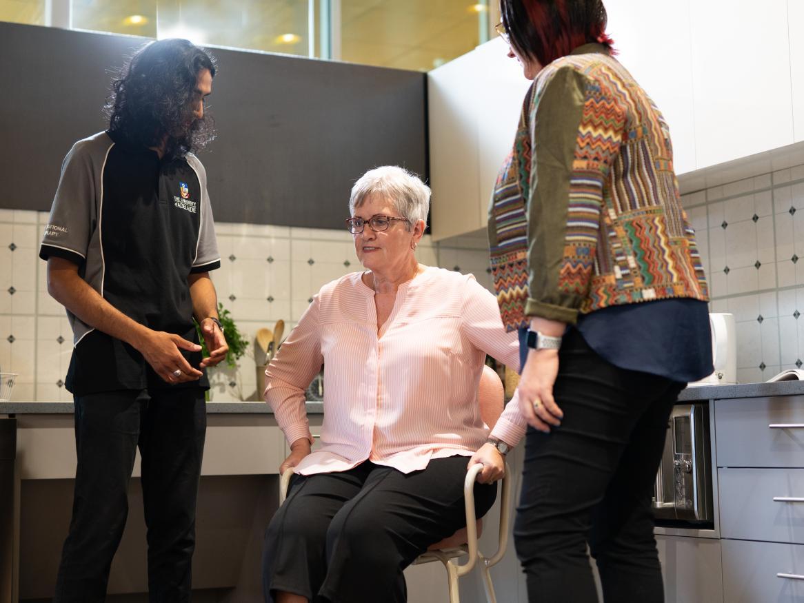 Occupational therapy student and teacher assisting older woman out of a chair in a kitchen