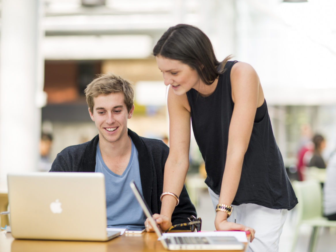 Student sitting in front of laptop with second student standing and leaning over to look at laptop